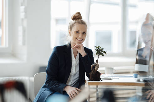 Smiling businesswoman sitting with plant on desk at office - JOSEF09100