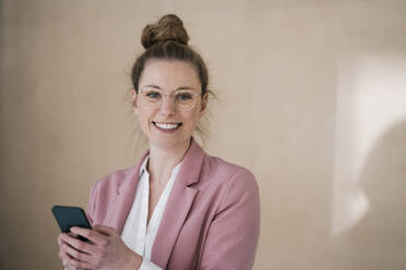 Happy young businesswoman holding smart phone in front of wall - JOSEF09103