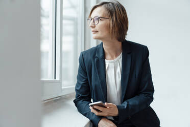 Smiling businesswoman with smart phone looking out through window - JOSEF09113