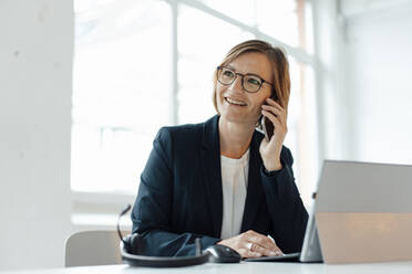 Lächelnde Geschäftsfrau, die am Schreibtisch im Büro sitzt und mit ihrem Smartphone spricht - JOSEF09119