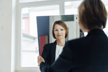 Businesswoman looking through mirror reflection at office - JOSEF09137