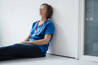 Tired female doctor leaning on wall relaxing at hospital corridor - JOSEF09148