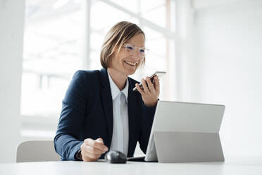 Happy businesswoman talking on mobile phone sitting at desk in office - JOSEF09151