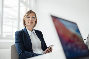 Businesswoman with mobile phone sitting at desk in office - JOSEF09153