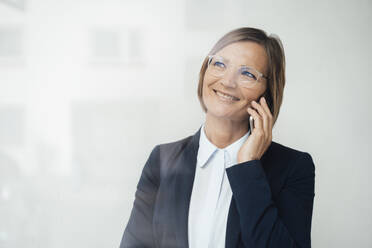 Glückliche Geschäftsfrau mit Blazer, die mit einem Smartphone telefoniert, gesehen durch ein Glasfenster - JOSEF09160
