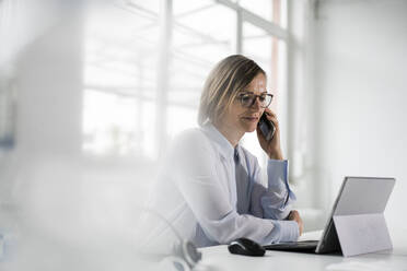 Businesswoman talking on mobile phone sitting at desk in office - JOSEF09174