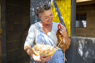 Smiling mature farm owner carrying hen and egg's bowl in front of chicken coop - ESTF00070