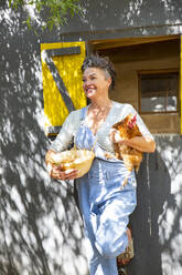 Smiling mature farmer holding hen and egg's bowl leaning on chicken coop - ESTF00072