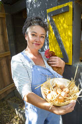 Smiling mature woman with hen and bowl of eggs standing in front of chicken coop - ESTF00077