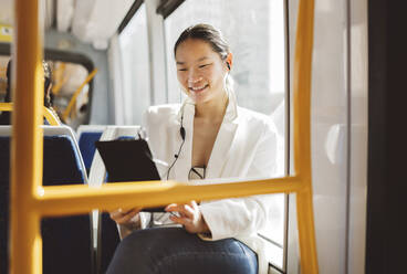 Smiling businesswoman holding tablet PC listening music through in-ear headphones in tram - JCCMF06178