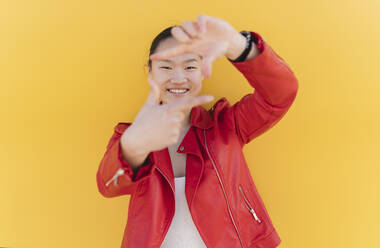 Smiling young woman making finger frame against yellow background - JCCMF06197