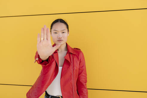 Junge Frau, die mit der Hand vor einer gelben Wand ein Stoppschild gebärdet - JCCMF06201