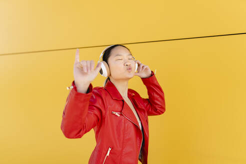Young woman listening music through wireless headphones showing Shaka sign in front of yellow wall - JCCMF06209