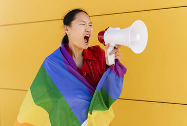 Eine in eine Regenbogenflagge gehüllte Frau schreit durch ein Megaphon an einer gelben Wand - JCCMF06215