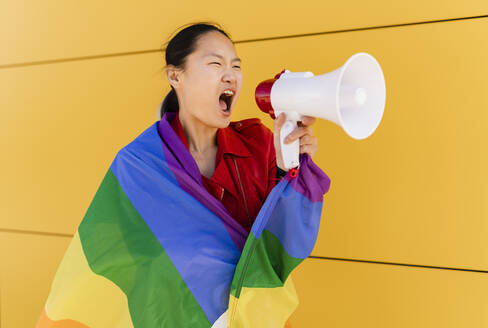 Eine in eine Regenbogenflagge gehüllte Frau schreit durch ein Megaphon an einer gelben Wand - JCCMF06215