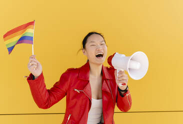 Happy woman holding rainbow flag shouting through megaphone in front of yellow wall - JCCMF06216