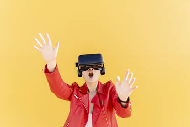 Young woman wearing virtual reality simulator gesturing against yellow background - JCCMF06217