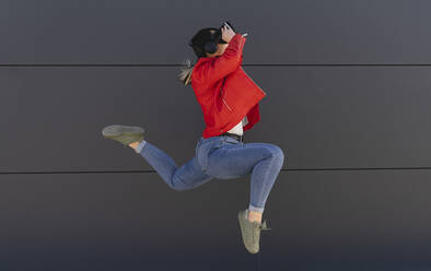 Playful woman wearing virtual reality simulator jumping in front of gray wall - JCCMF06220