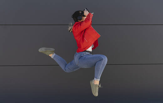 Verspielte Frau im Virtual-Reality-Simulator, die vor einer grauen Wand springt - JCCMF06220