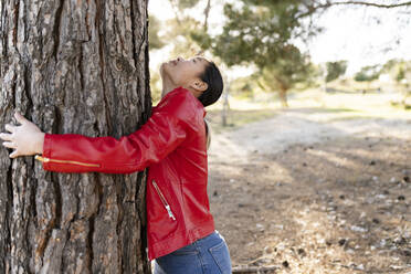 Young woman wearing red jacket embracing tree in forest - JCCMF06221