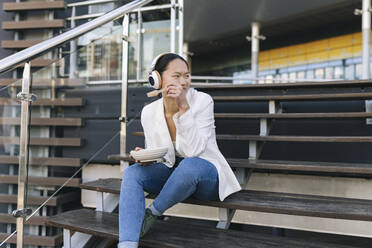 Smiling businesswoman holding bowl listening music through wireless headphones sitting on steps - JCCMF06238