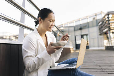 Glückliche Geschäftsfrau isst Salat und sitzt mit Laptop - JCCMF06241