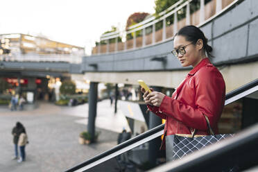 Junge Frau mit Einkaufstasche und Smartphone, die auf einer Rolltreppe nach unten fährt - JCCMF06246