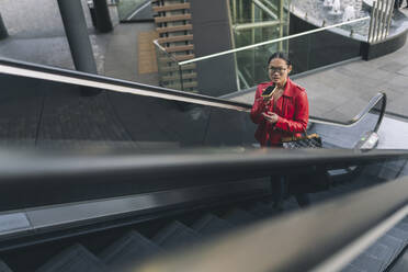 Junge Frau, die auf einer Rolltreppe über den Lautsprecher ihres Mobiltelefons spricht - JCCMF06247