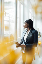 Geschäftsfrau hält Smartphone und schaut durch ein Fenster im Büro - JOSEF09224