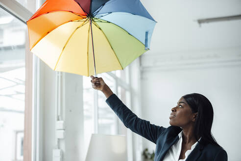 Businesswoman holding colorful umbrella in office - JOSEF09228