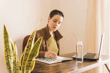 Freelancer with laptop and book sitting at table at office - MGRF00646