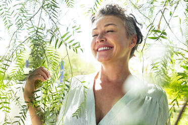 Lächelnde Frau mit geschlossenen Augen inmitten von Zweigen im Garten stehend - ESTF00095