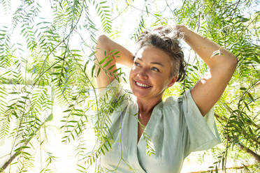 Lächelnde reife Frau inmitten von Zweigen an einem sonnigen Tag - ESTF00097