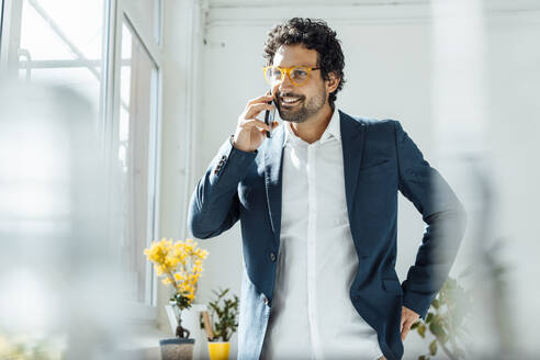 Lächelnder Geschäftsmann mit Brille, der im Büro mit seinem Smartphone telefoniert - JOSEF09252