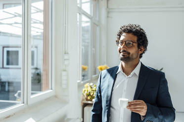 Lächelnder Geschäftsmann mit Kaffeetasse, der durch ein Fenster im Büro schaut - JOSEF09263