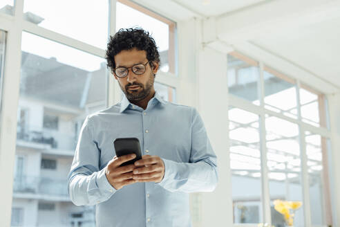 Businessman using smart phone standing in front of window - JOSEF09267