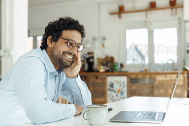 Happy businessman wearing eyeglasses looking at laptop in office - JOSEF09273