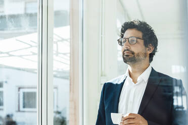Thoughtful businessman holding coffee cup looking through window in office - JOSEF09297