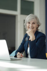 Happy businesswoman with hand on chin sitting at desk - JOSEF09304