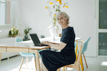 Happy businesswoman with laptop sitting on chair at desk in office - JOSEF09316