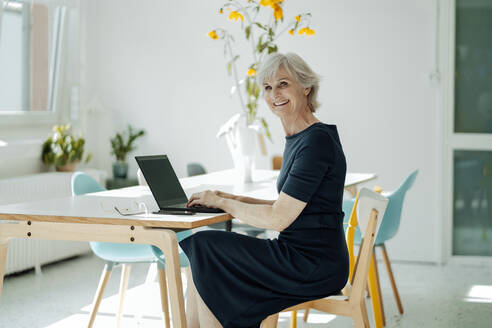 Glückliche Geschäftsfrau mit Laptop auf einem Stuhl am Schreibtisch im Büro sitzend - JOSEF09316