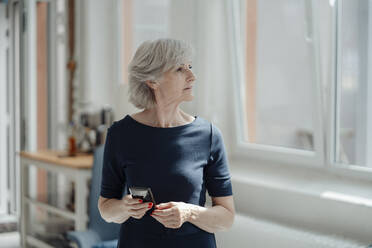 Senior businesswoman with gray hair holding smart phone in office - JOSEF09317