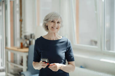 Smiling senior businesswoman with mobile phone standing in office - JOSEF09318