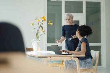 Senior businesswoman discussing with colleague sitting at desk in office - JOSEF09326