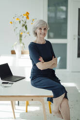 Smiling businesswoman with arms crossed sitting on desk in office - JOSEF09327
