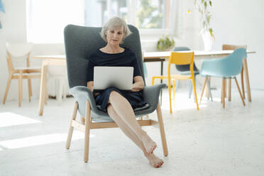 Senior businesswoman using laptop sitting in armchair in office - JOSEF09338