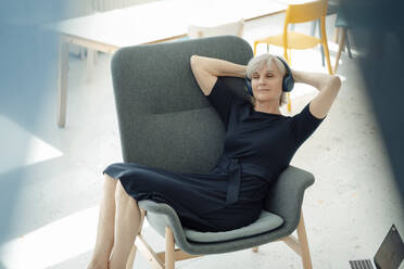 Ältere Geschäftsfrau mit drahtlosen Kopfhörern, die sich mit den Händen hinter dem Kopf in einem Sessel im Büro entspannt - JOSEF09339