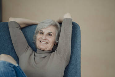 Happy senior woman sitting on chair in front of wall - JOSEF09365