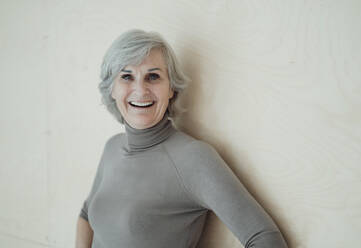 Happy senior woman with gray hair standing in front of wall - JOSEF09369