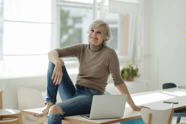 Smiling businesswoman with laptop sitting on desk in office - JOSEF09374